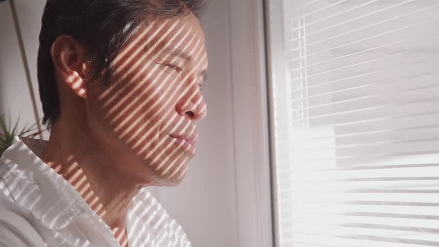 A man looks out a window as the sun shines through the blinds casting shadows and lines on his face