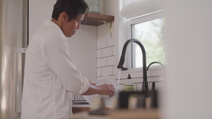 Man standing at kitchen sink next to a window washing a plate