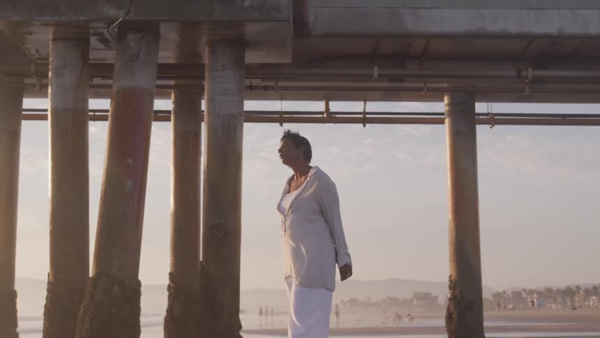 A woman stands on the beach with her hands behind her back as she holds her face up to the wind