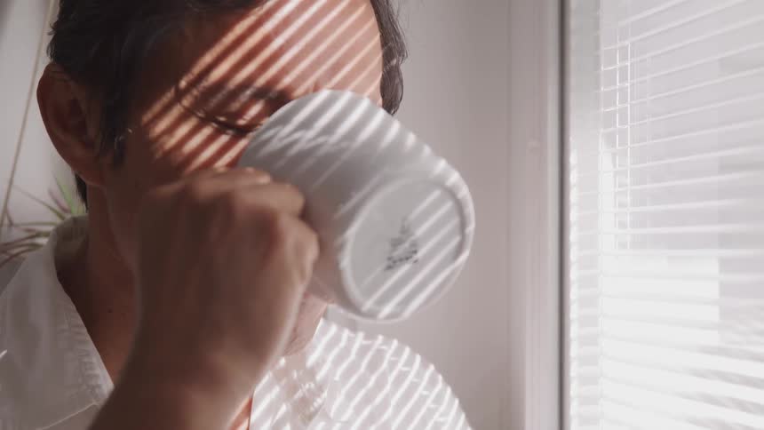 Asian man drinks a cup of coffee and looks out the window in a room