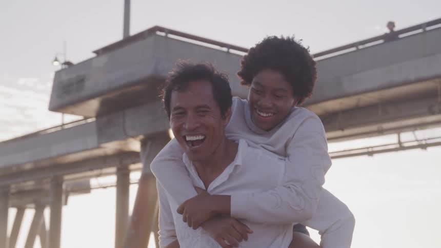 A man twirls and laughs as he gives his wife a piggy back ride near the pier