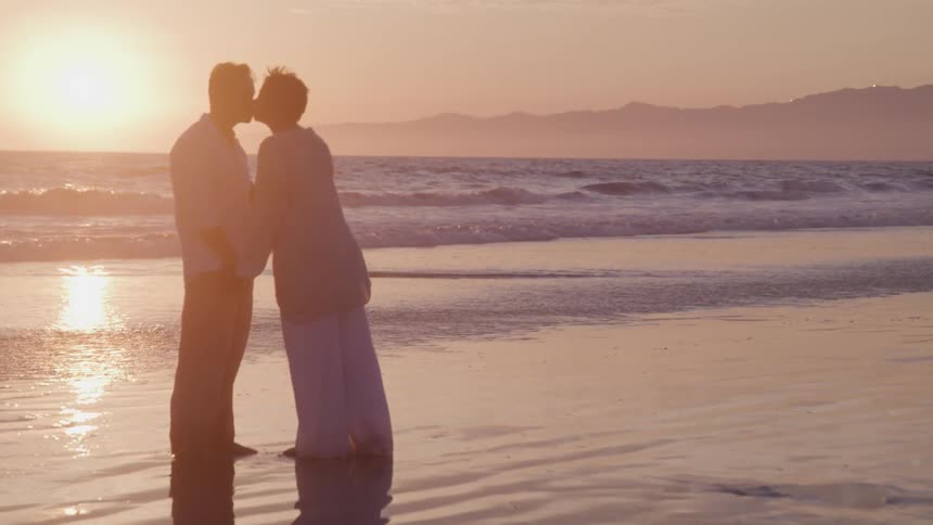 Asian man and black woman kiss on the beach at sunset next to the ocean