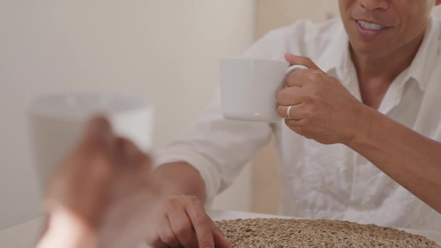 Asian man sitting at the table holds a cup of coffee and talks to his friend in a room