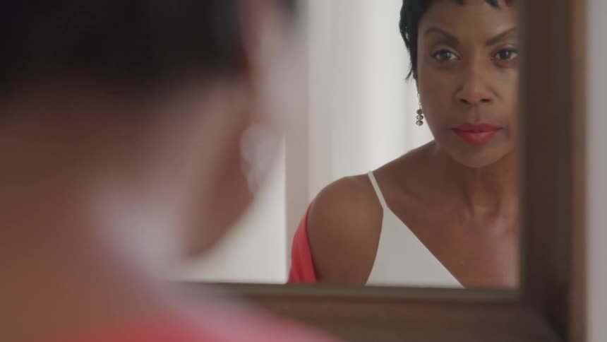 A black woman wearing a white dress looks up at her reflection in a mirror
