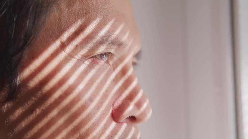 A man stares of of window blinds with the sunlight hitting his face