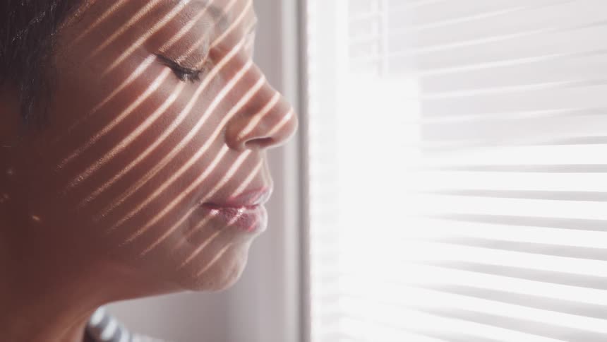 Woman stands with eyes closed next to open blinds