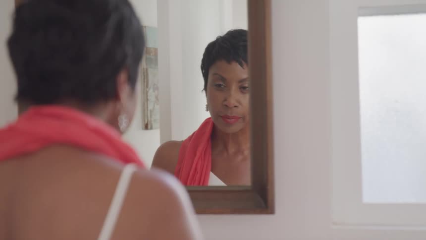 An older black woman stands in front of a mirror and stares at herself