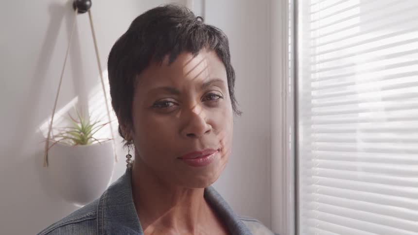 A short haired black woman looking into the camera while standing near a window indoors