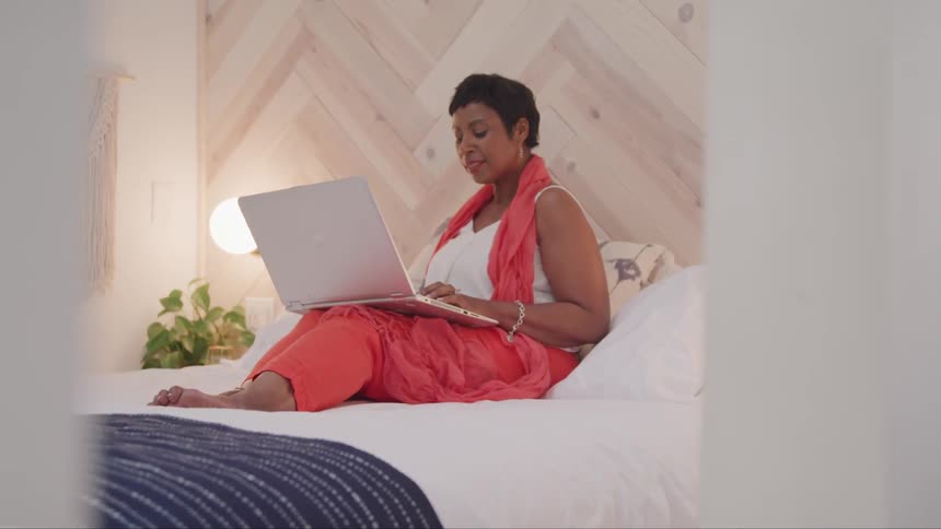 Black woman sitting on the bed uses a laptop in the bedroom