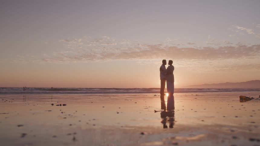 Asian woman and black woman stands on the beach and hold each other next to the ocean at sunset