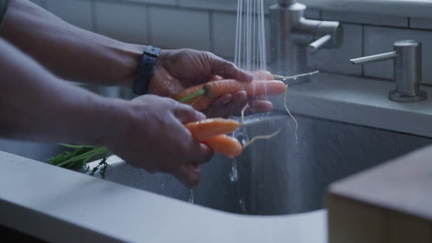 A person holds and washes carrots under the spray in the kitchen sink