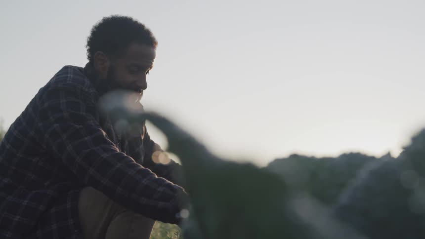 A black man kneels and fiddles with an object outdoors