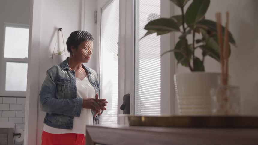 A woman leans on a glass door with her hands together in front of her as the sun shines through the blinds