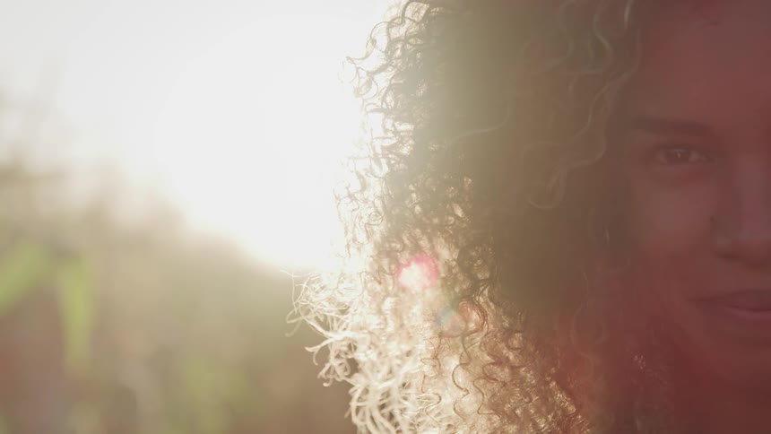 A young black woman raising her eyes to look into the camera as the sun shines through her hair