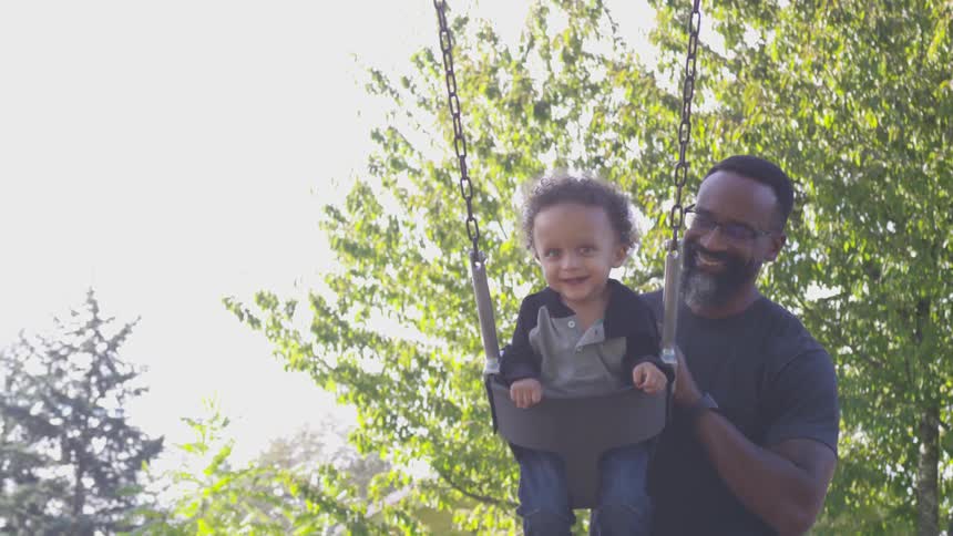 A father smiles while pushing his young son on an outdoor swing
