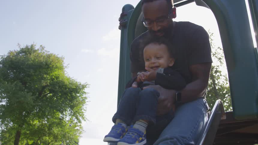 A man holds a toddler going down a park slide together