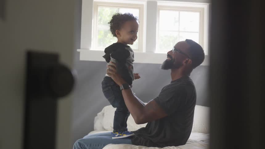 Black man sitting on the bed holds toddler boy in the bedroom