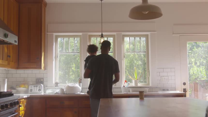 Black man holding toddler boy stands in front of the kitchen counter in the kitchen