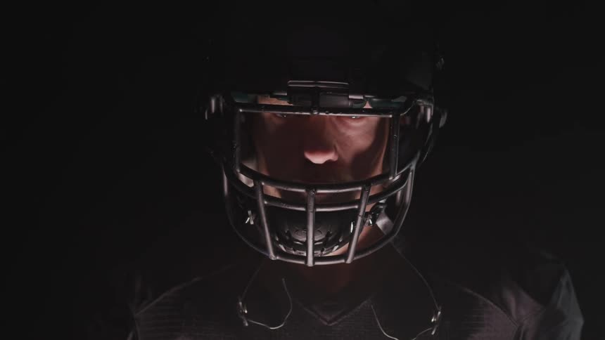 A man wearing a football helmet and black jersey is slowly illuminated by the light