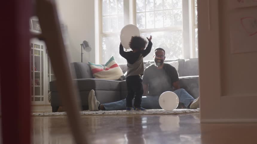 Black man sitting on the ground plays with toddler boy with white balloon in the living room