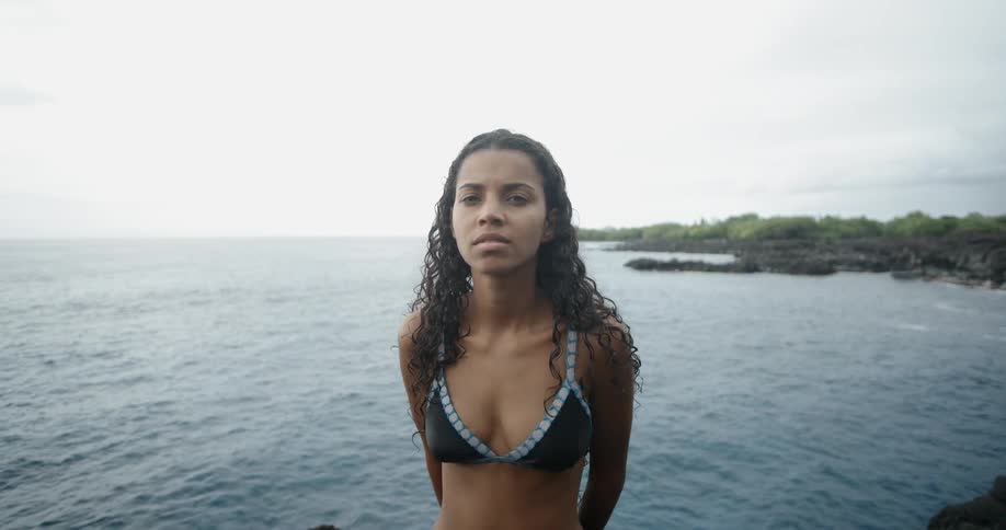 A woman stands on a rocky ledge on the coast wearing a bikini