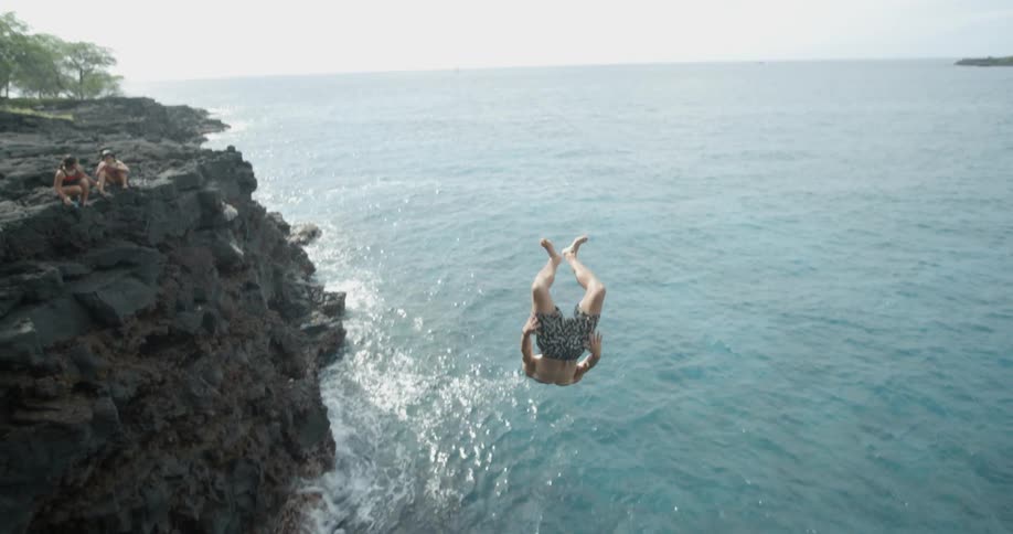 A man does a back flip dive off of a cliff as people watch