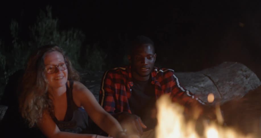 Man and woman roast food over bonfire outdoors at night