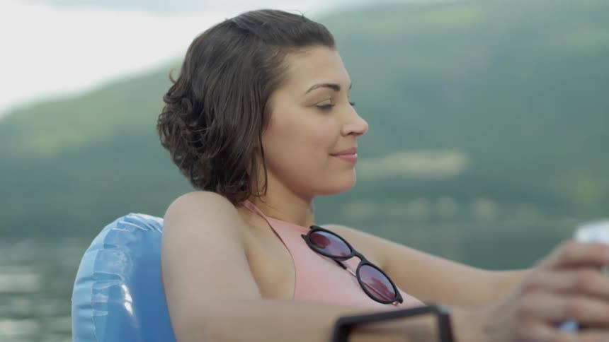 Woman drinks beer on a floating pool chair in a lake near mountain