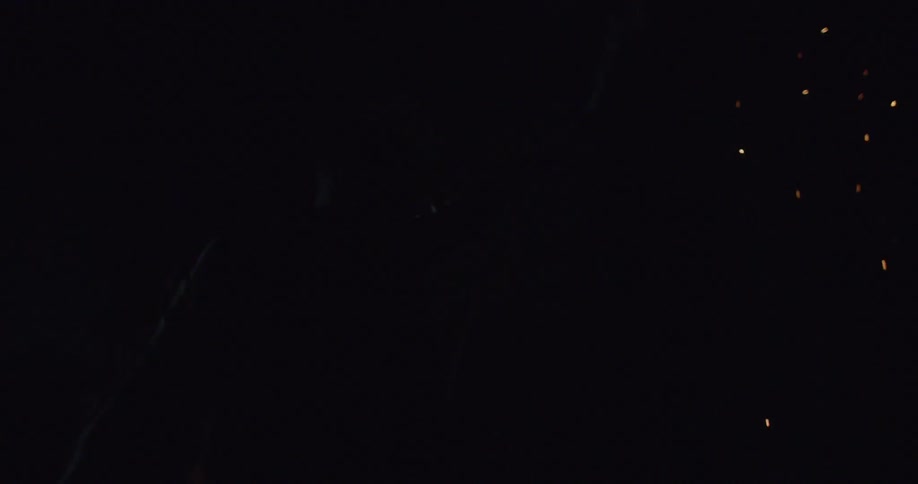 A black man standing in a woodland holding a roman candle as it fires sparks into the sky