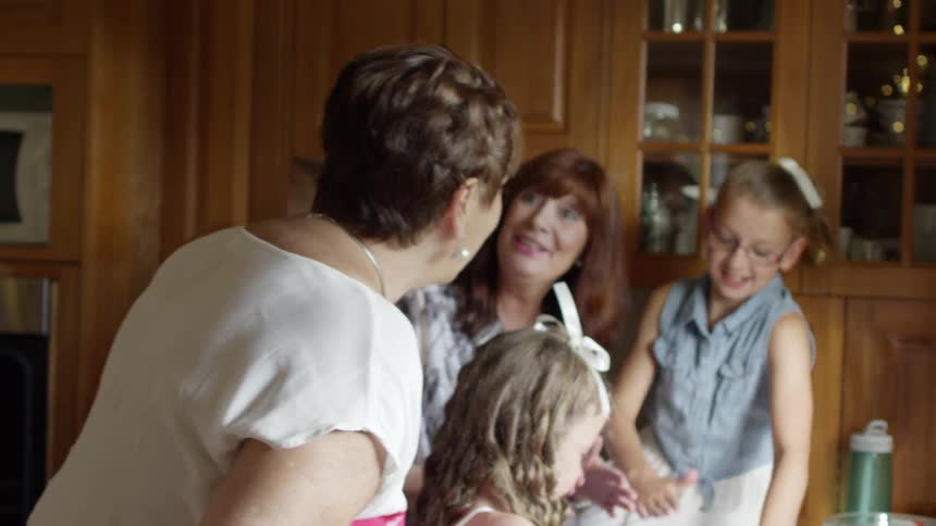 A woman and two young girls preparing food in the kitchen when another woman walks in and speaks to them