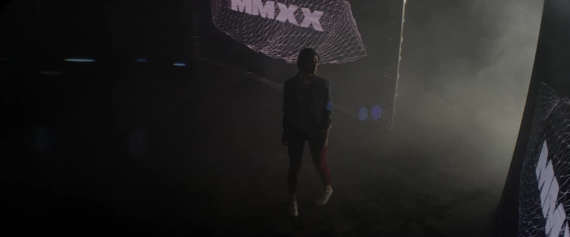 A woman walks from a smoky corridor into a dark room with abstract imagery and text projected onto the walls