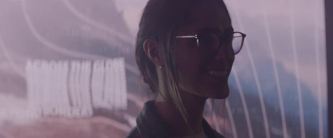 A woman wearing glasses stands before a screen with abstract images and wavy lines projected onto it