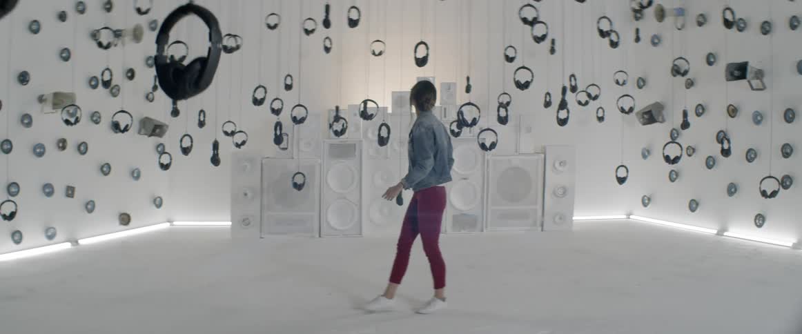 A woman stands in an art installation with speakers mounted on walls and headphones hanging from the ceiling