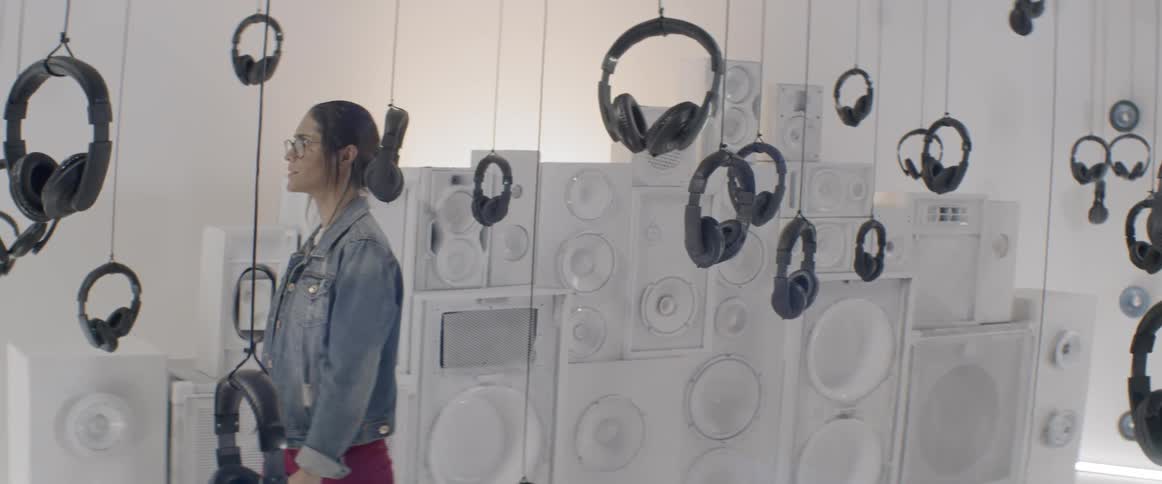 A woman walking through a room examining the many sets of headphones hanging from the ceiling on strings