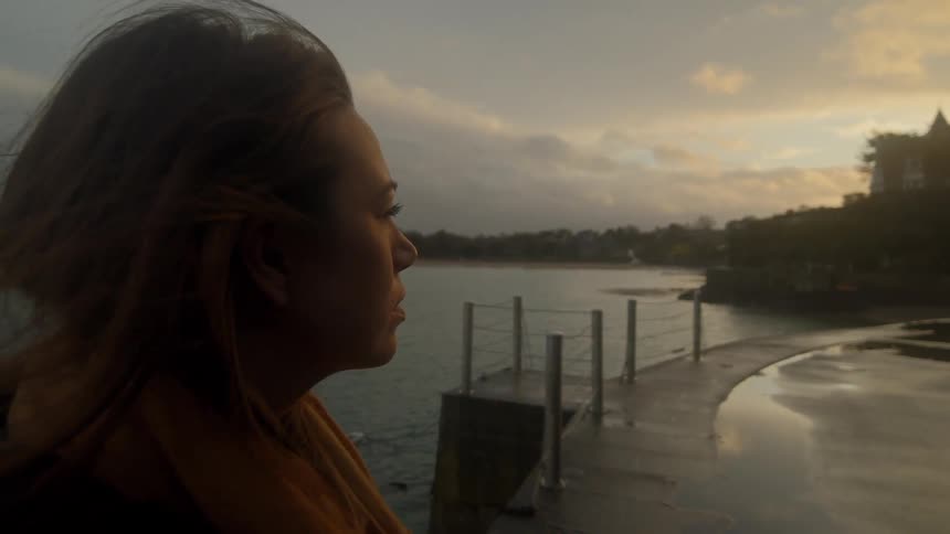 A woman stands on a rain covered pier on a lake during sunset