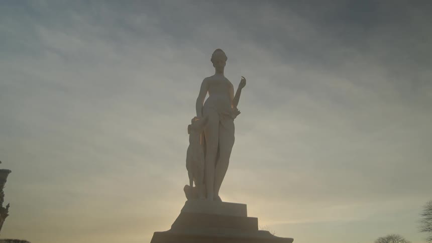 A tall statue of a female figure standing with a dog is atop a pedestal under a cloudy sky