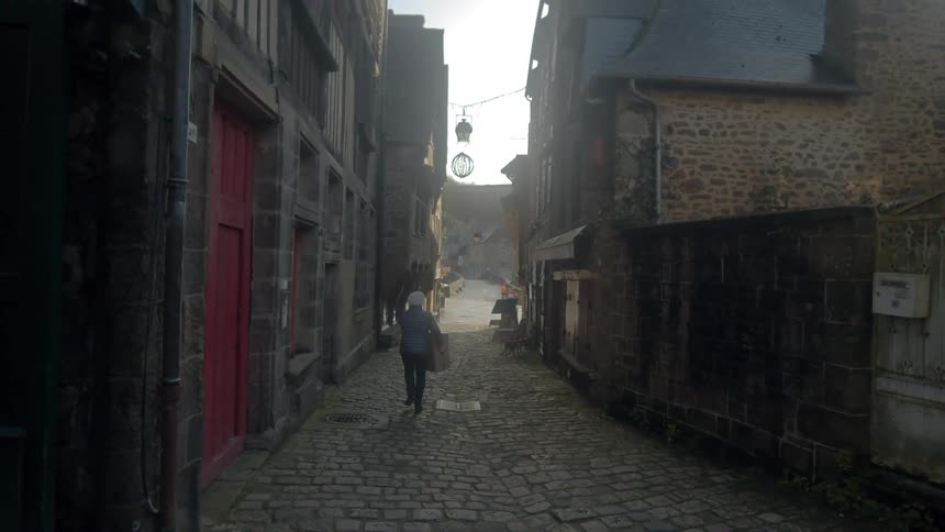 An elderly carries a box down a brick street lined with buildings