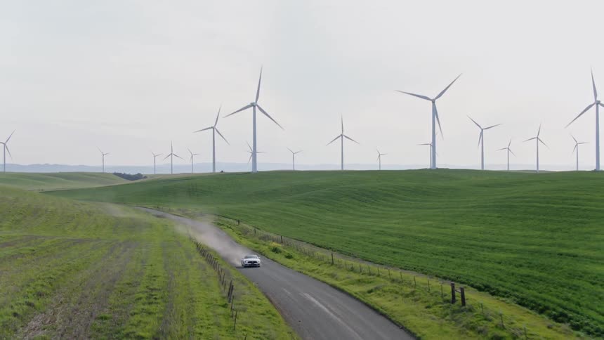 Car kicks up dust as it drives along road next to wind farm