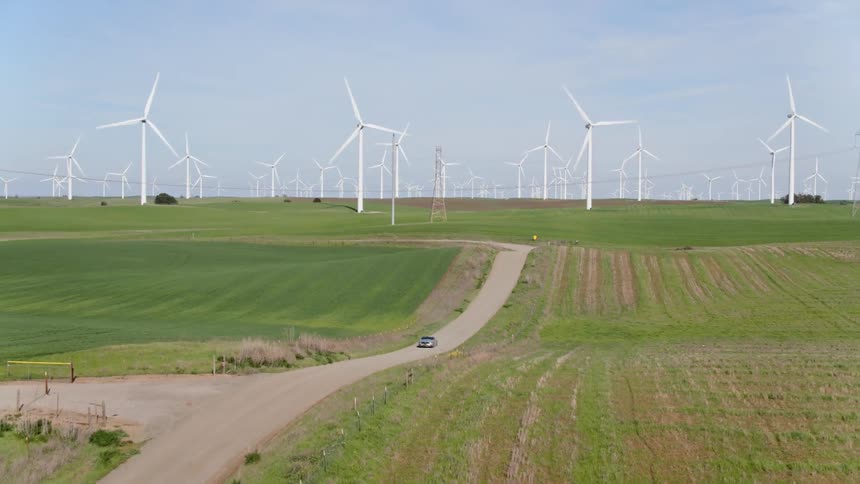 A car travels on a road away from a wind farm with large white turbines