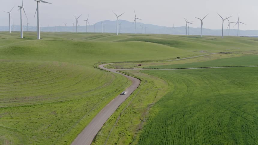 A car travels down a narrow road that winds through a wind farm