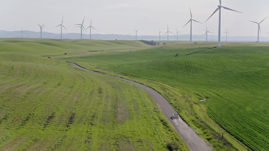 A vehicle drives a lone roadway beside large wind turbines