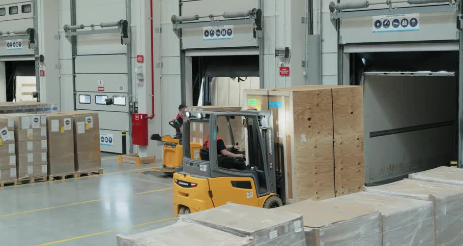 A man drives a full forklift backwards out of a tractor trailer in a warehouse