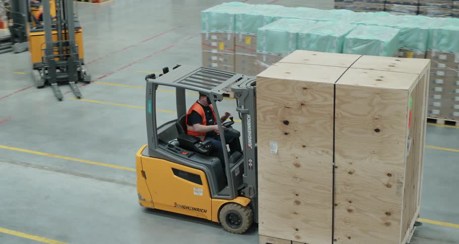A forklift driver is moving a large box through the warehouse while other forklifts drive empty