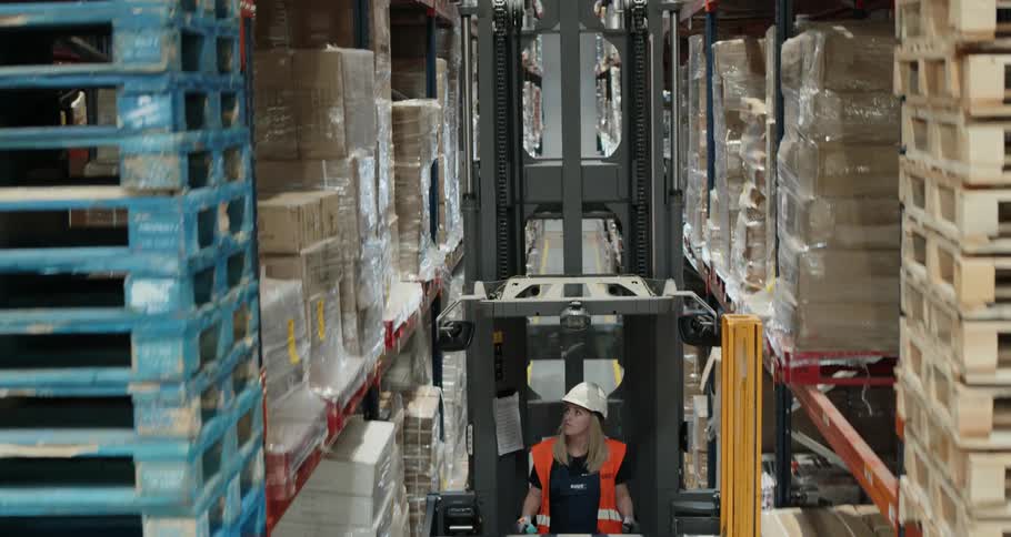 A woman operates a narrow aisle forklift and rises up to the high shelves in a warehouse