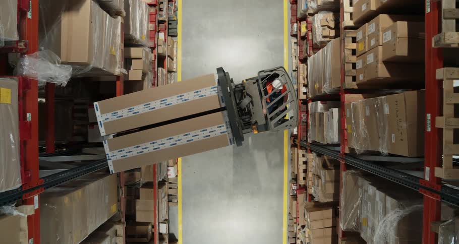 Within a warehouse full of tall shelves a person driving a forklift removes a pair of large boxes from a high shelf