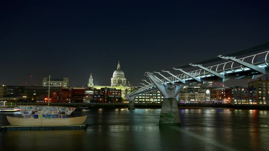 Several boats pass under a bridge at night near a metropolitan area