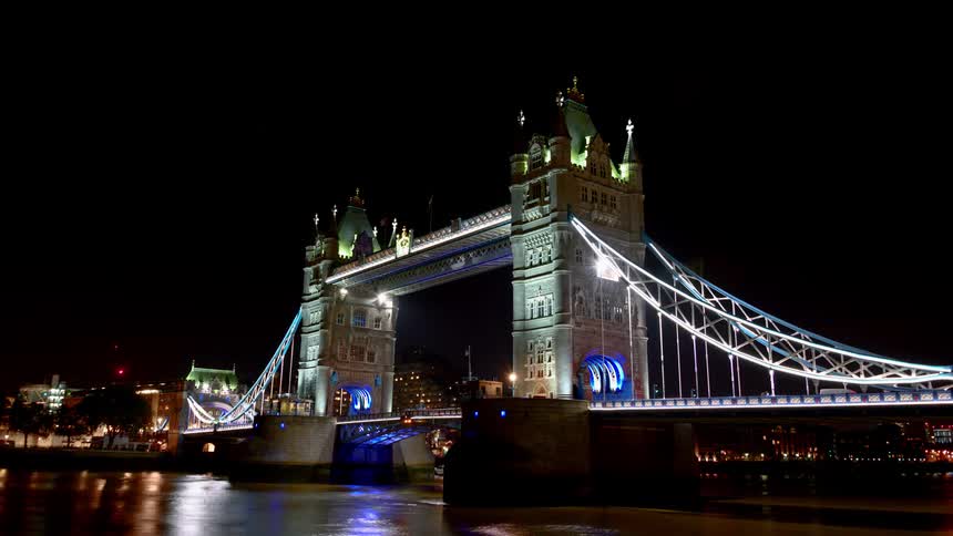 The london bridge is brightly illuminated at nighttime as vehicles pass over it and boats cruise on the river beneath it