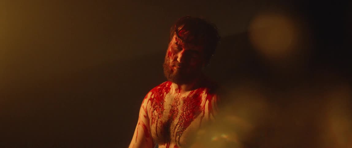 A bloody caucasian man wearing a crown of thrones slowly closing his eyes and lowering his head