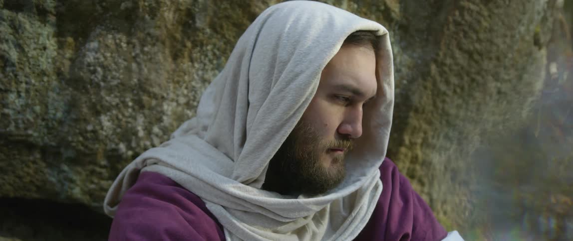 Man wearing robes and headcovering rests near stone wall