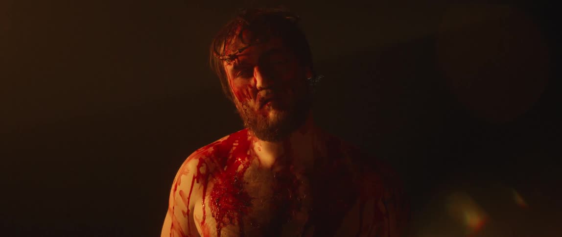 A bearded and shirtless man covered in bloods stands still as he struggles to keep his head up